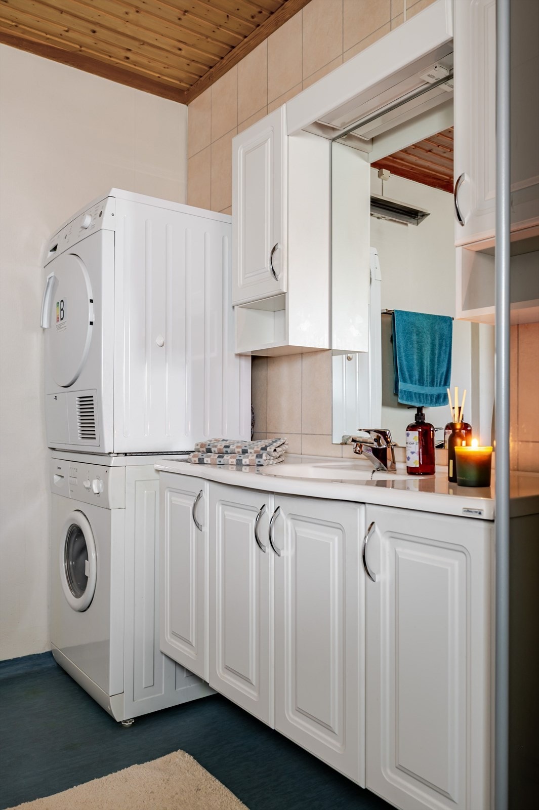 Lys baderomsinnredning med heldekkende servant, speil med belysning og vegghengte skap over. Baderommet er også utstyrt med dusjhjørne, toalett og opplegg for vaskemaskin/tørketrommel. Galleribilde