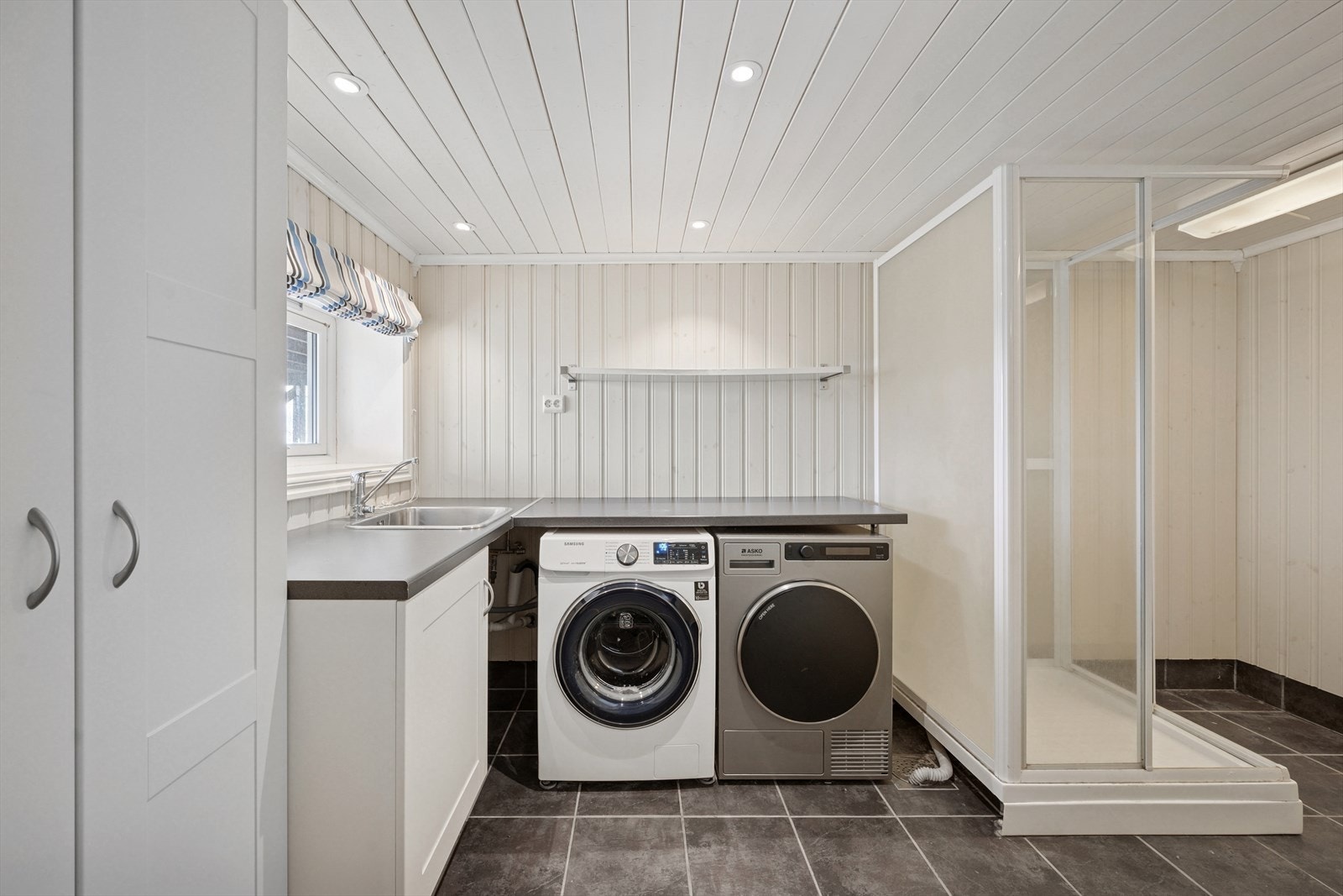 Malt panel på vegger. Fliser på gulv. Dusjkabinett, utslagsvask i benk og opplegg for vaskemaskin. El. avtrekksvifte på yttervegg. Varme i gulv. Galleribilde