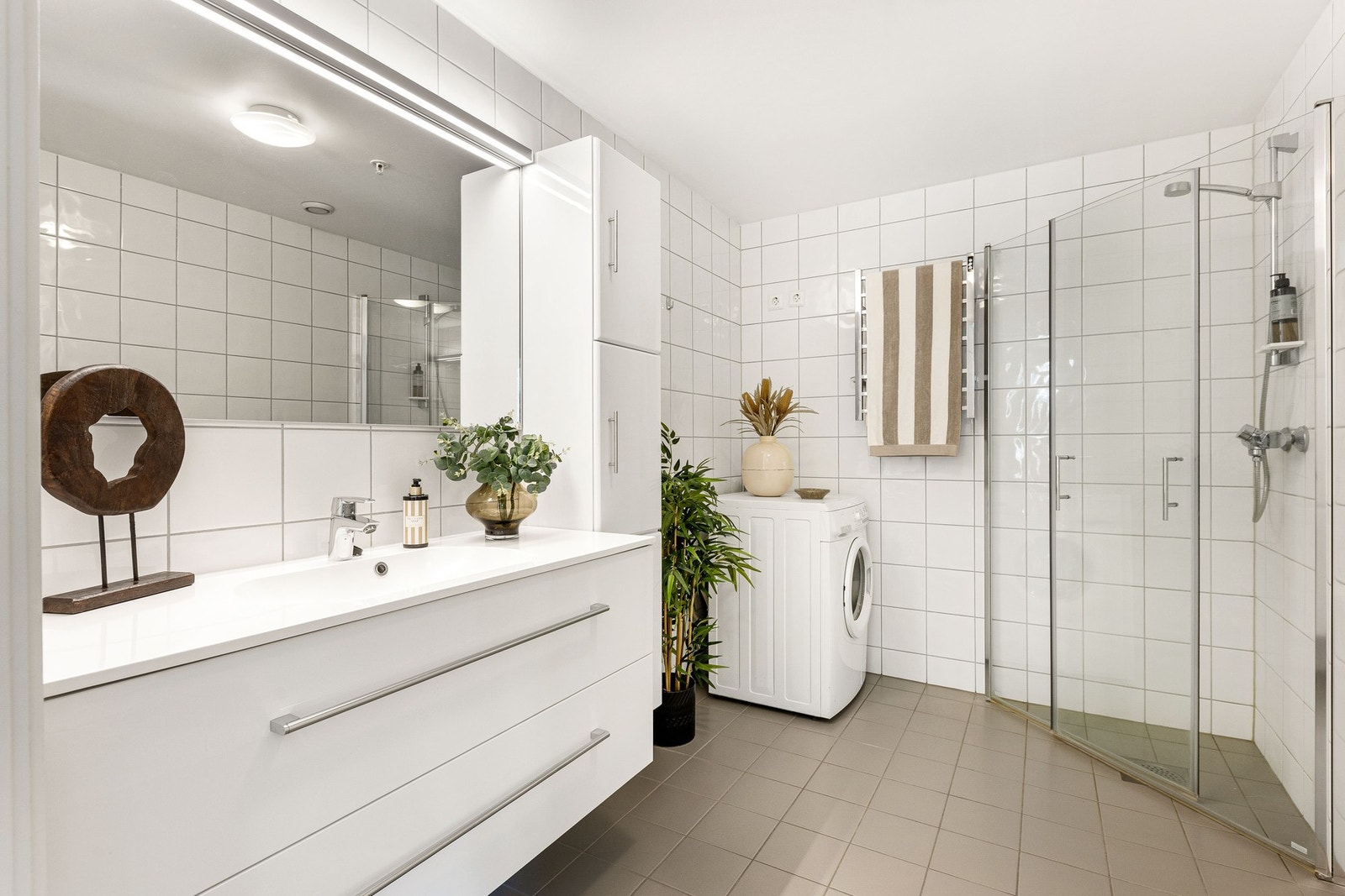 Lyst og moderne bad med dusjkabinett, plass til vaskemaskin og tørketrommel. Flislagte vegger og gulv gir et stilrent uttrykk. Galleribilde