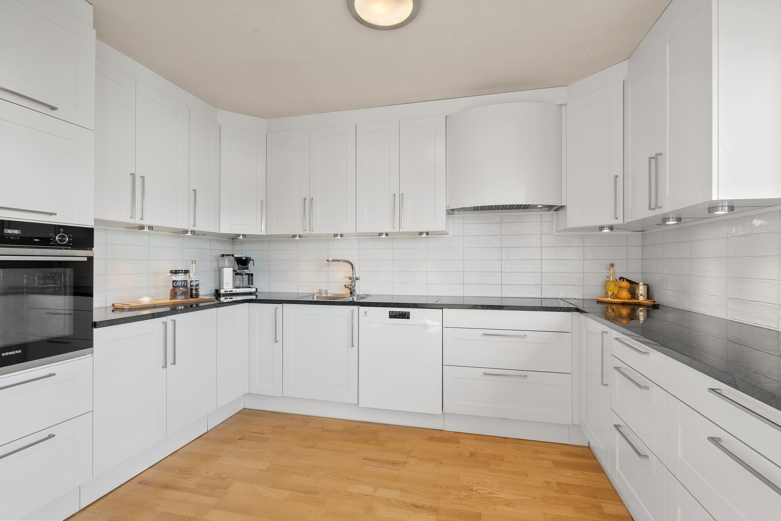 Pent og tidløst kjøkken fra 2011 med hvite profilerte fronter og påkostet benkeplate av stein/granitt. Fliser på vegg over benk. Lys under overskap. Galleribilde