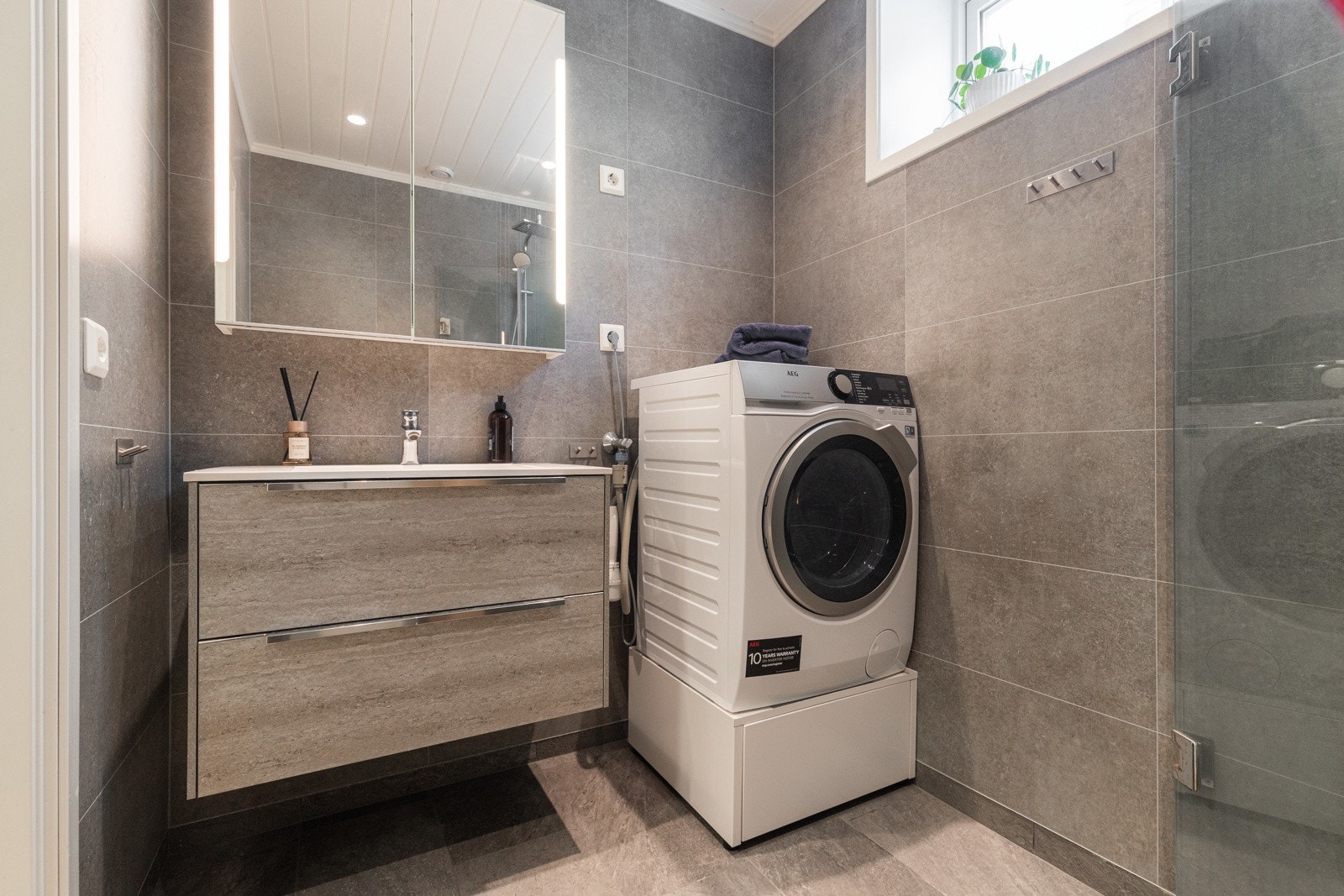 Badet er stilfullt innredet med speil med integrert belysning, nedfelt servant med tilhørende skuffer, klosett, praktisk dusjnisje og opplegg for vaskemaskin. Galleribilde