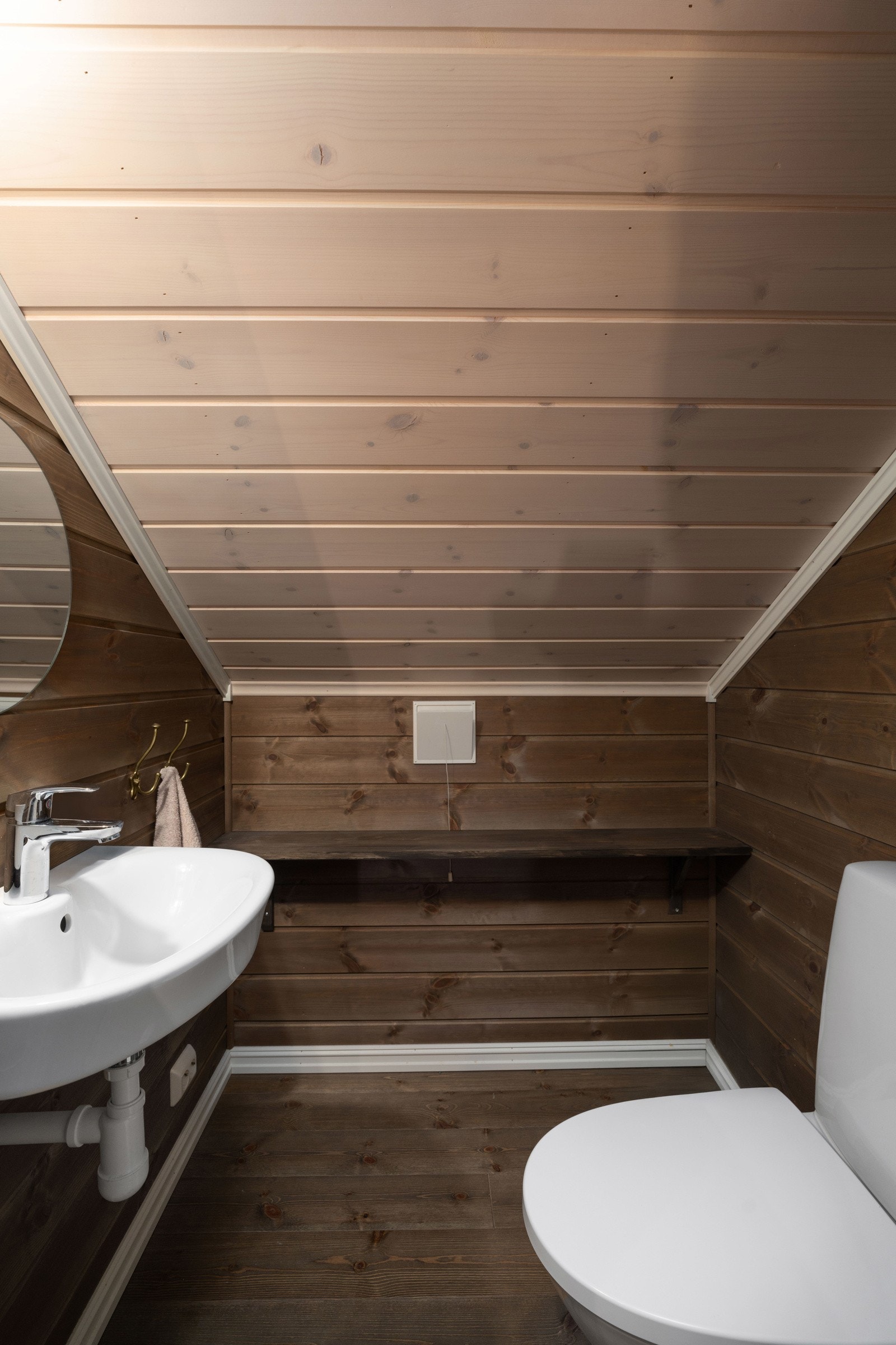 Toalettrommet har mørkbeiset furugulv og tilsvarende farge på veggene, avbrutt av hvitmalte lister og hvitbeiset panel i skråhimling. Galleribilde