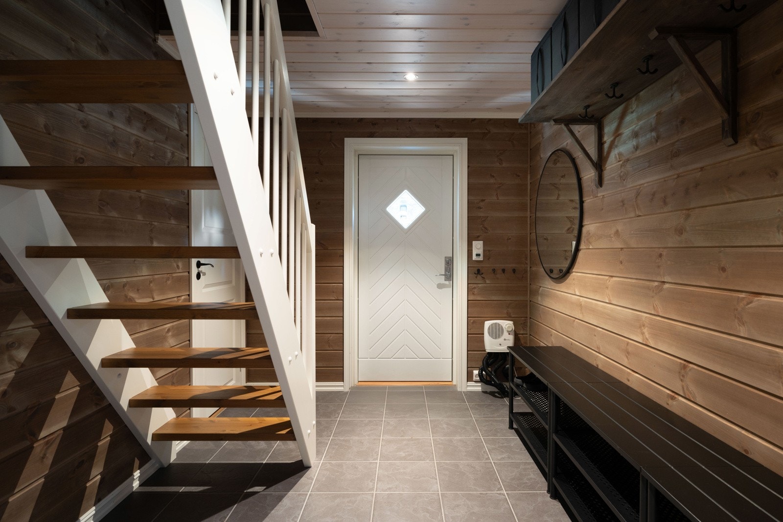 Entreen strekker seg ut i lengde og har god plass til turtøy og sko. En åpen, rett trapp fører opp til hems, og det er dør inn til en bod. På gulvet er det grå fliser med gulvvarme. Galleribilde