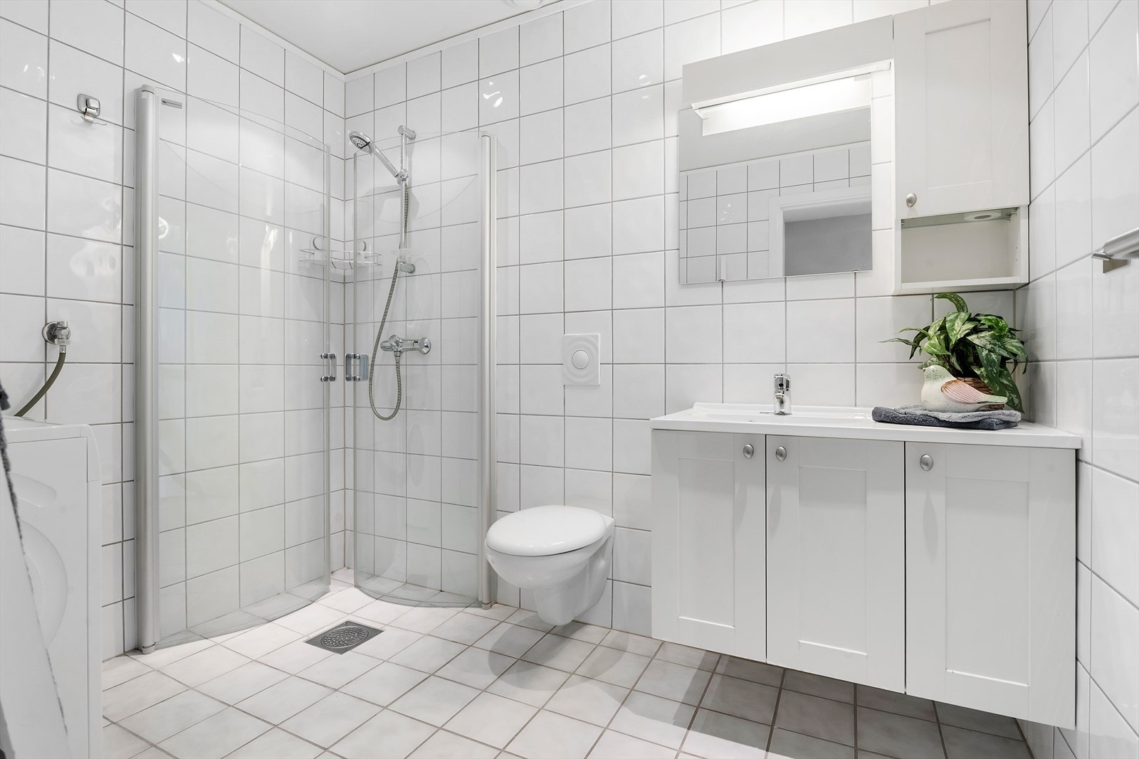 Flislagt bad/vaskerom med vegghent toalett, dusjddører, innredning med servant og opplegg for vaskemaskin. Galleribilde