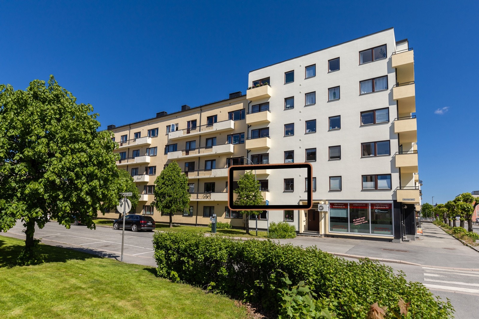 Leilighetsbygg med 6 etasjer, balkonger, forretning i første etasje. Grøntområde og gateparkering foran bygget. Galleribilde