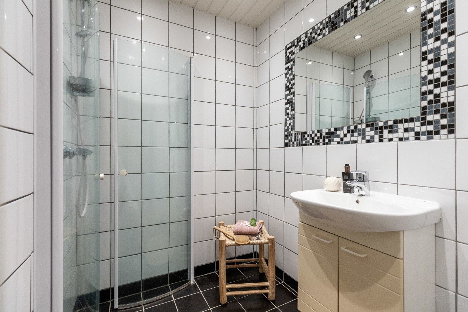 Lyst bad med dusjhjørne, speil med mosaikkfliser, og servant med underskap. Veggene er flislagt. Galleribilde