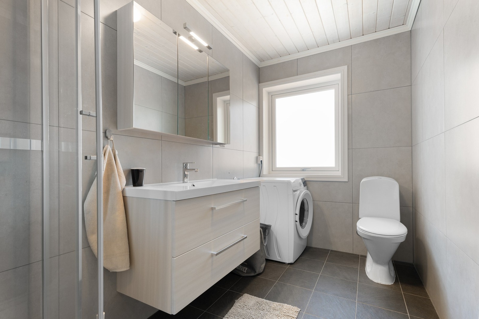 Baderom med dusjkabinett, toalett, innredning med servant og opplegg til vaskemaskin Galleribilde