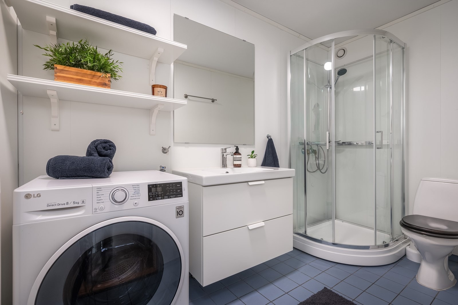 Lyst bad med servant med skuffer, speil, klosett, dusjkabinett og opplegg for vaskemaskin. Galleribilde