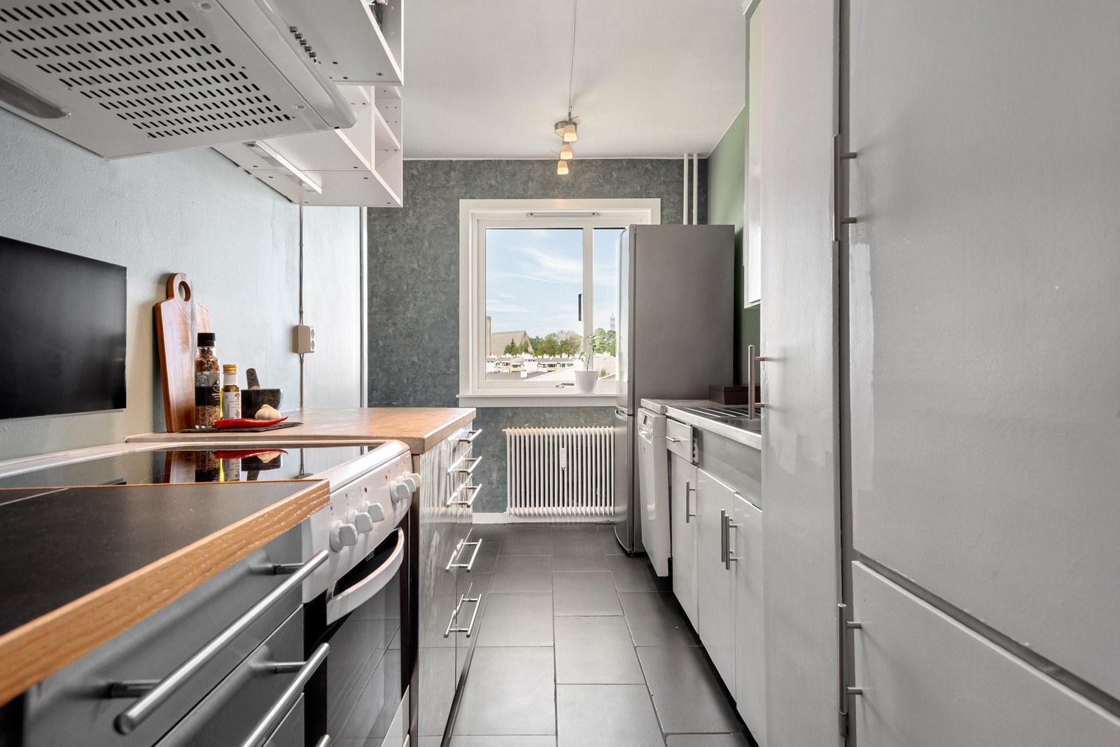 Rommet byr på godt med plass til innredning og oppbevaring, og dagens løsning består av glatte fronter, benkeplate, frittstående kjøleskap samt stekeovn med koketopp. Galleribilde