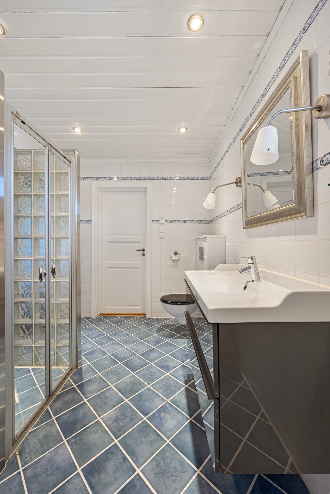 Badet har en stor dusjnisje i glassbyggerstein, veggmontert toalett og en moderne servantinnredning i mørk høyglans. Galleribilde