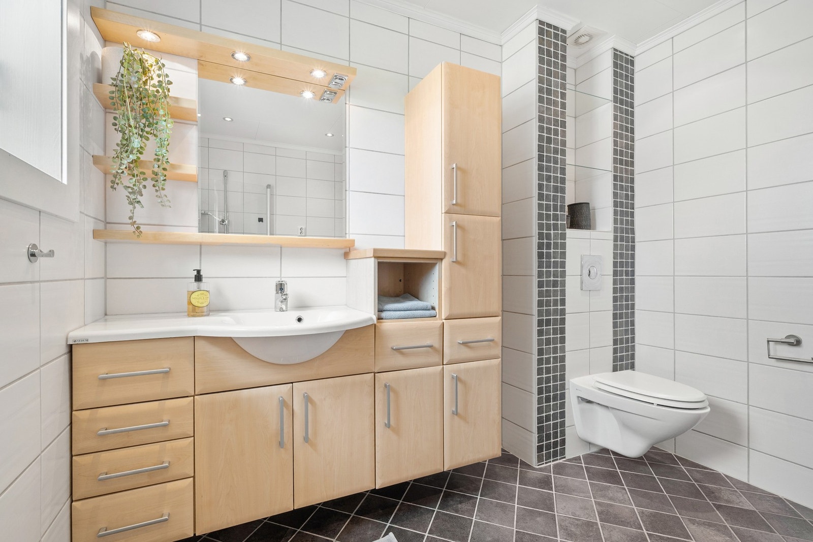 Pent flislagt bad med vegghengt toalett, servantskap, dusjhjørne og kran og avløp til vaskemaskin. Galleribilde