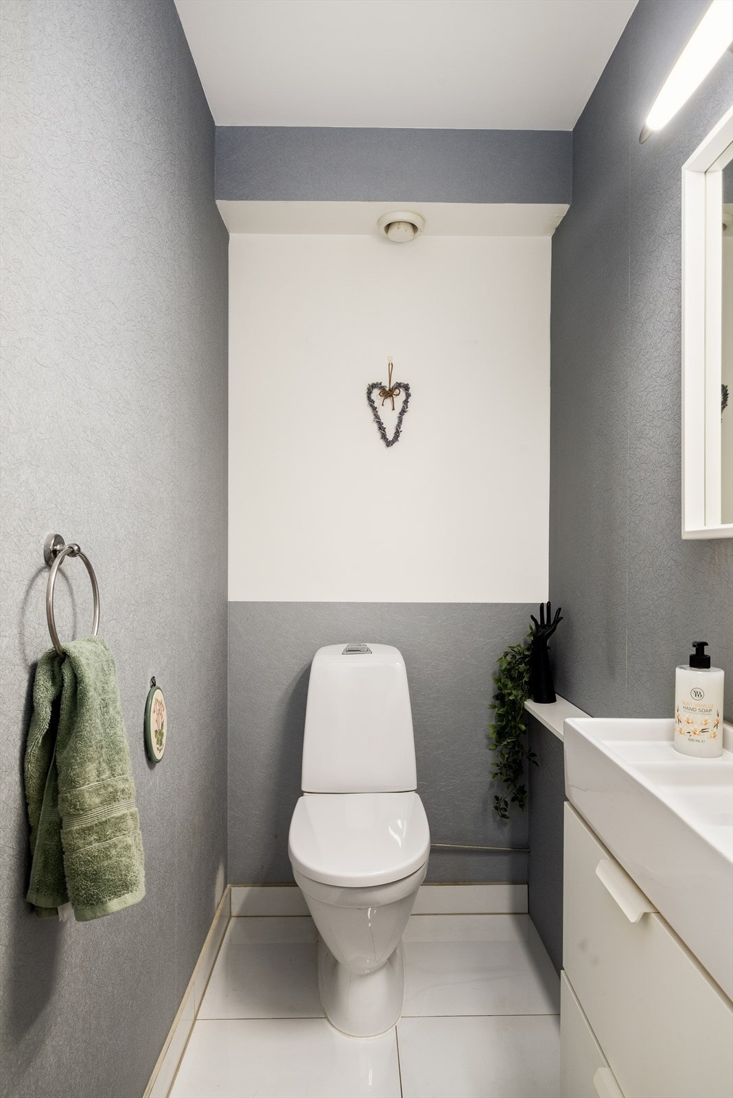 Toalettrom med flis på gulv og tapet på vegger. Tak har malt betong. Innredet med klosett, servant i innredning og speil. Galleribilde