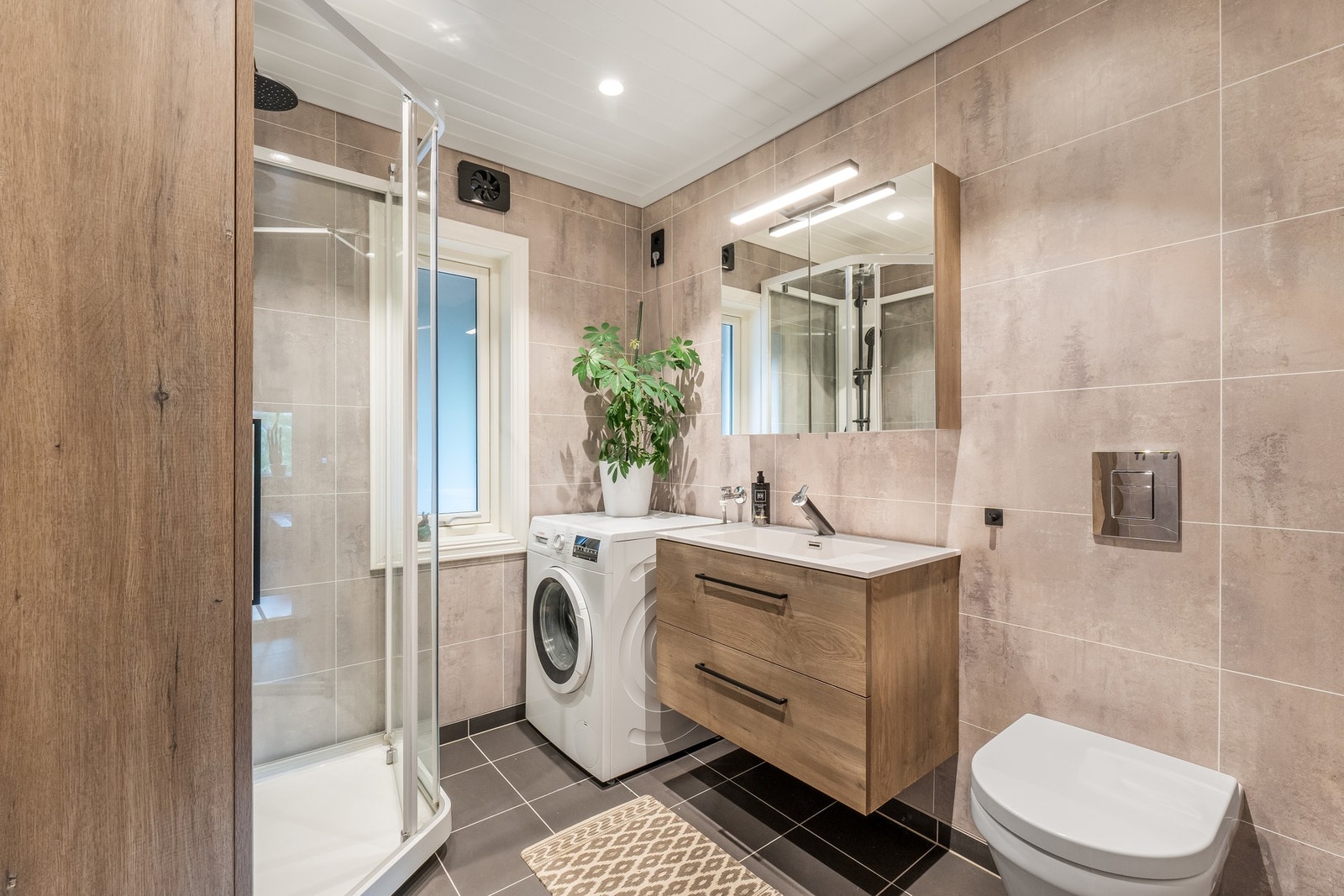 Badet i første etasje kombinerer funksjonene bad og vaskerom, med flislagt gulv, baderomsplater på veggene og panel i taket. Galleribilde