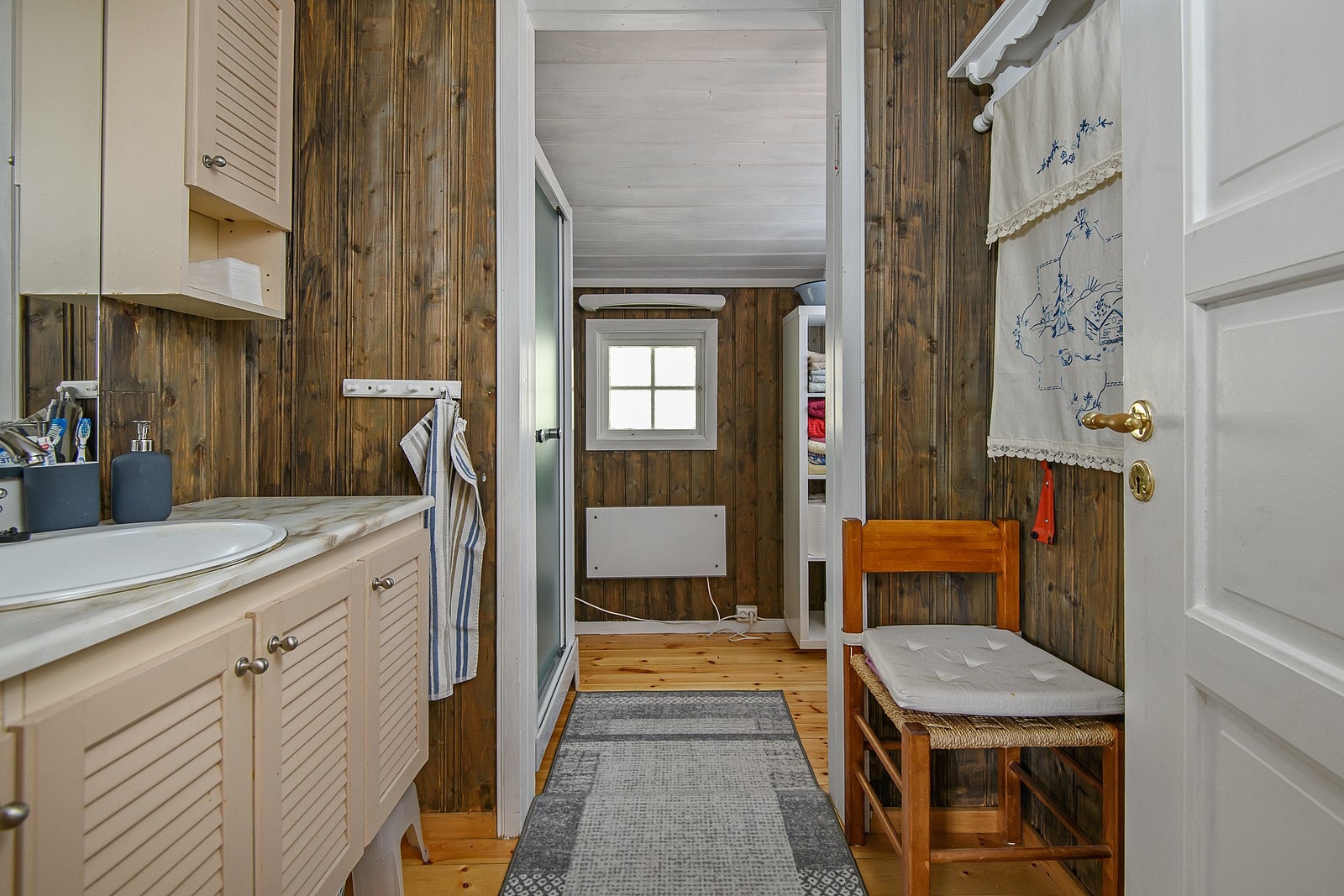 Baderom med tregulv og panelvegger. Innredningen består av servant med underskap og dusjkabinett. Det er panelovn på veggen. Galleribilde