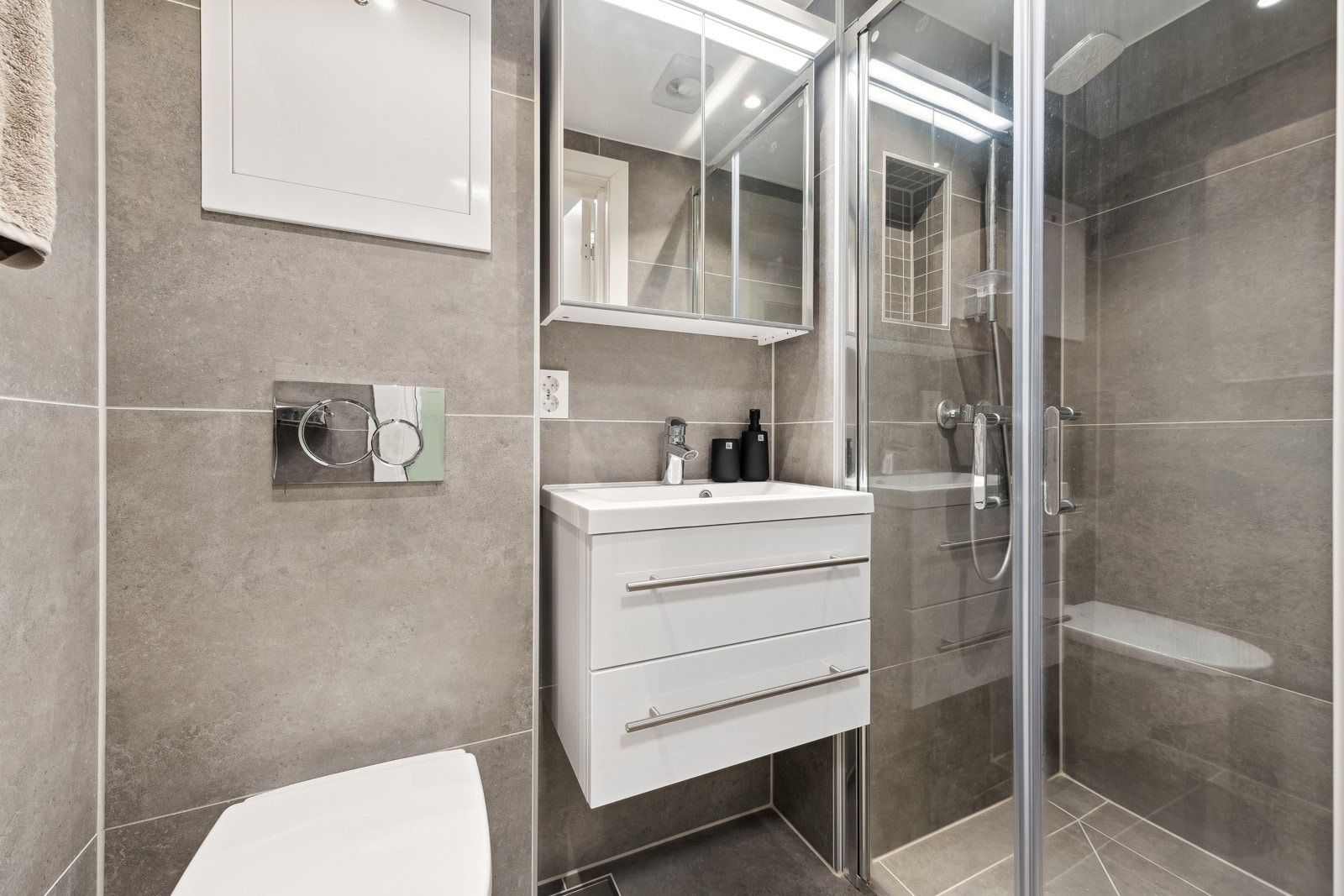 Badet er utstyrt med baderomsinnredning med servant med underskap (skuffeseksjon), speilskap med belysning, veggmontert toalett og dusjvegger/hjørne. Galleribilde