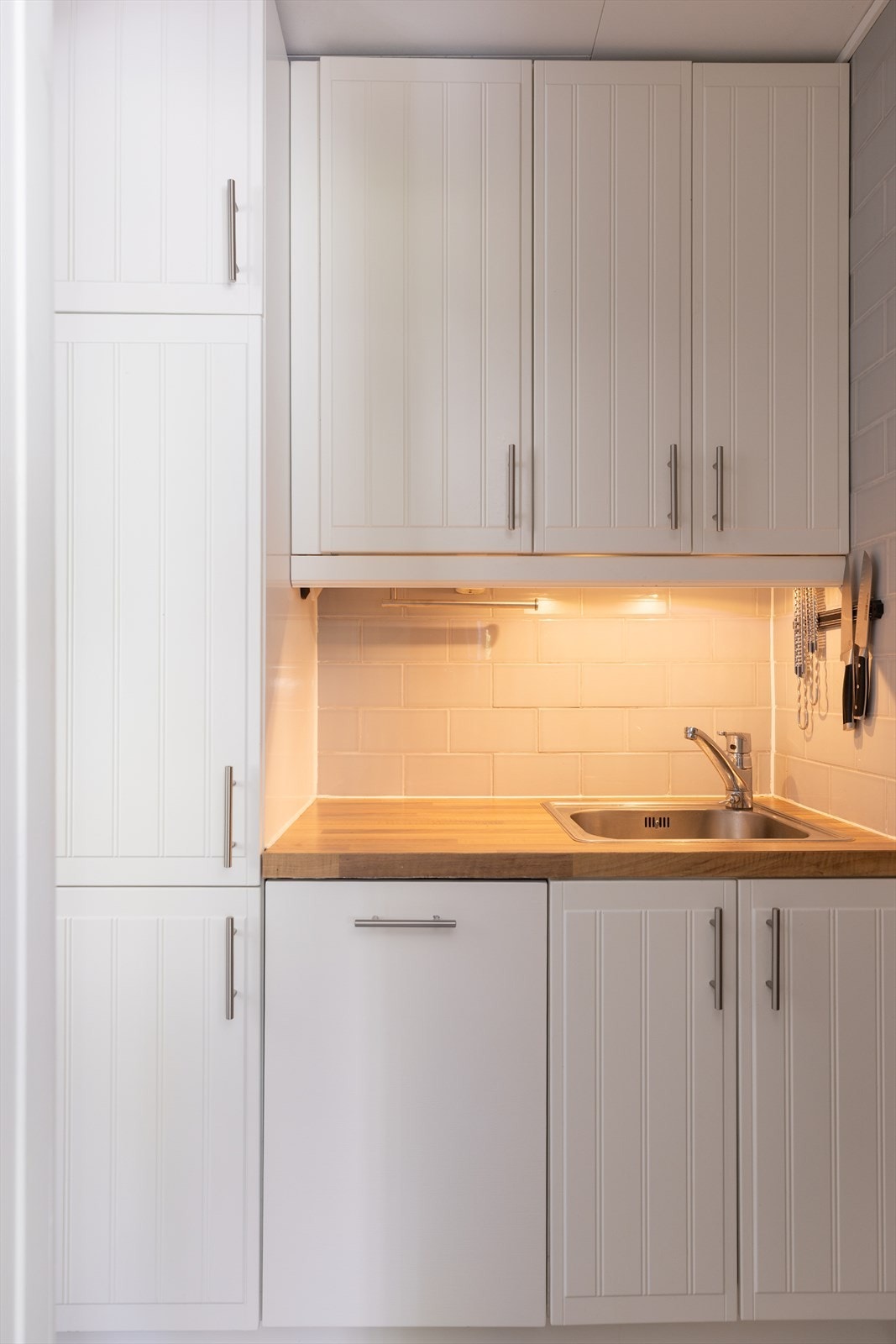 Kjøkkeninnredning med hvite profilerte fronter, laminat benkeplate, oppvaskkum i stål, fliser over benk. Galleribilde
