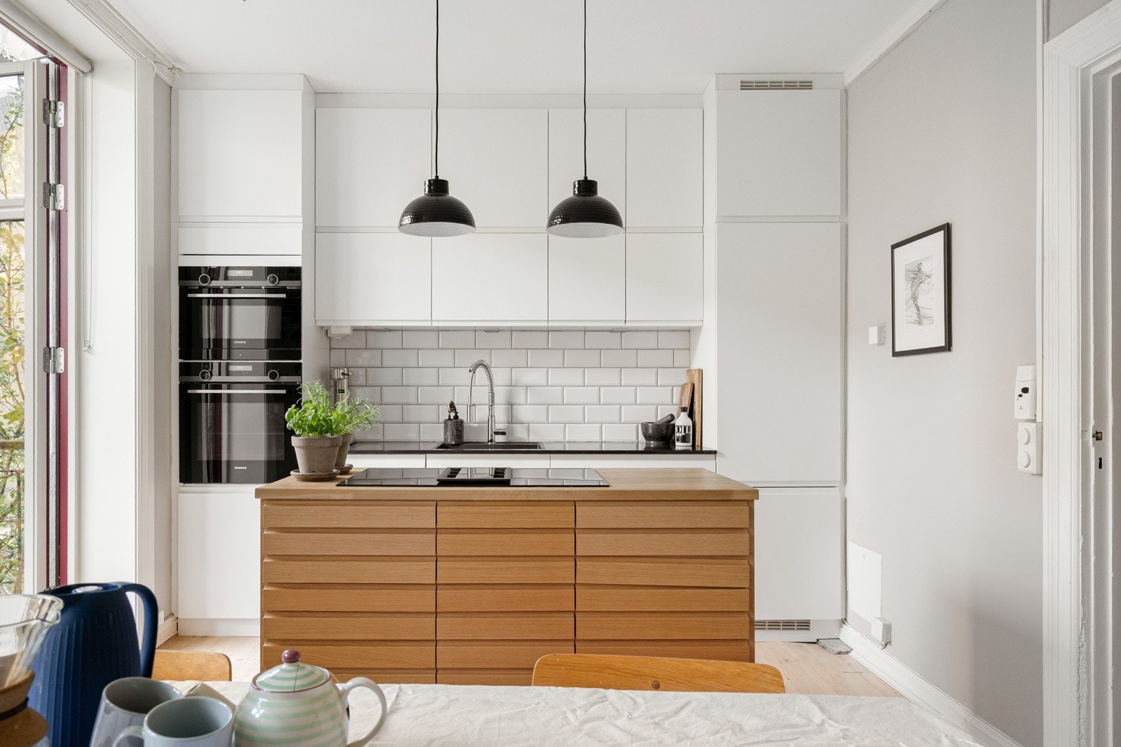 Innbydende kjøkken fra KVIK, nytt i 2019 med glatte, lyse fronter og benkeplate i stein med nedfelt kum i kompositt. Over benken er det fliser på vegg og belysning under overskapene som gir et funksjonelt og delikat uttrykk. Galleribilde