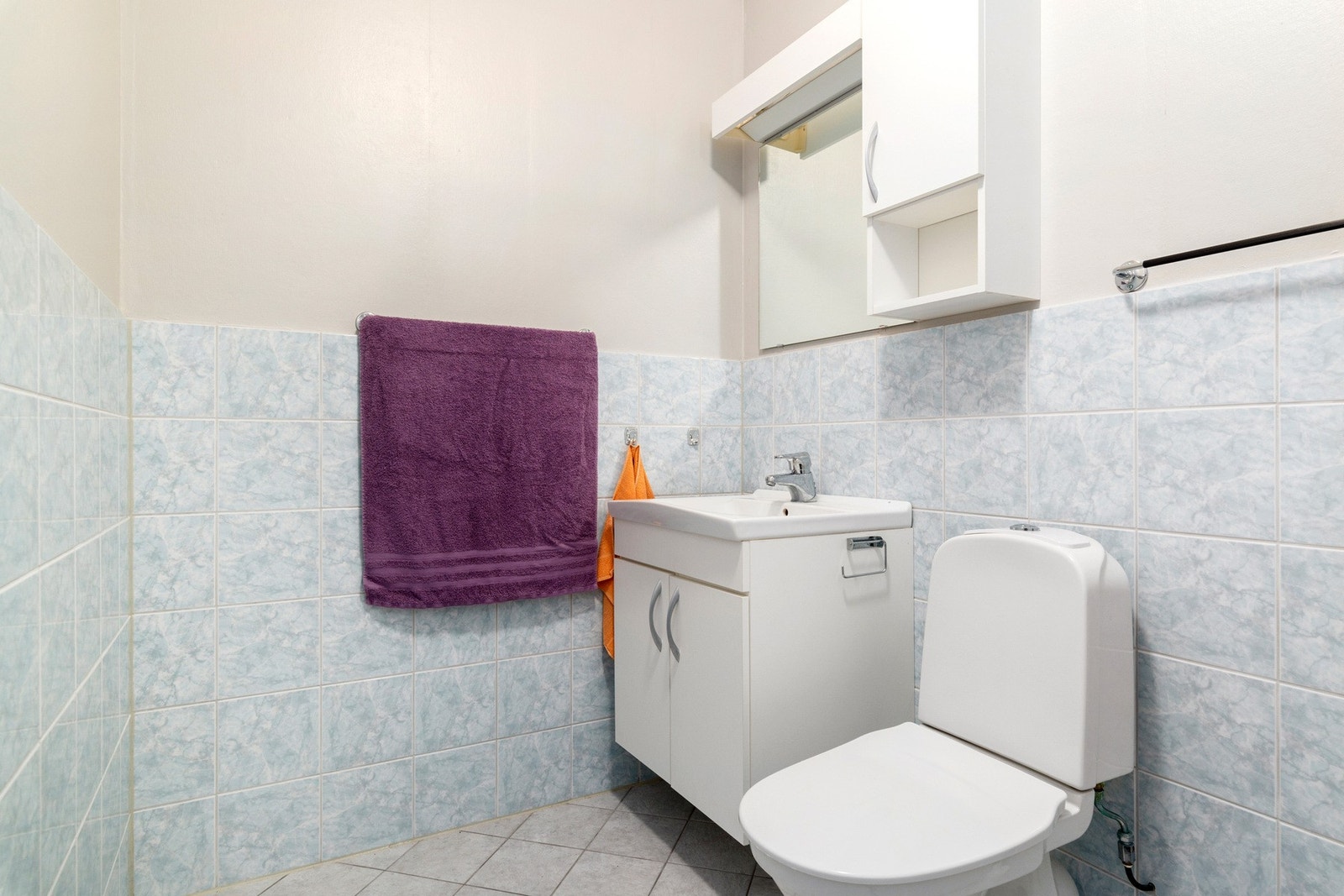 Badet har flislagte gulv og delvis på veggflater, gulvmontert wc og en enkel servantinnredning. Galleribilde