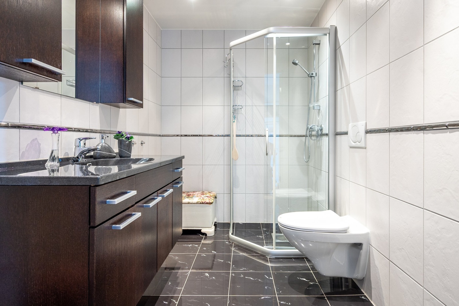 Flislagt bad innredet med vegghengt servantinnredning, speil med belysning og skaper, dusjhjørne og vegghengt toalett. Galleribilde