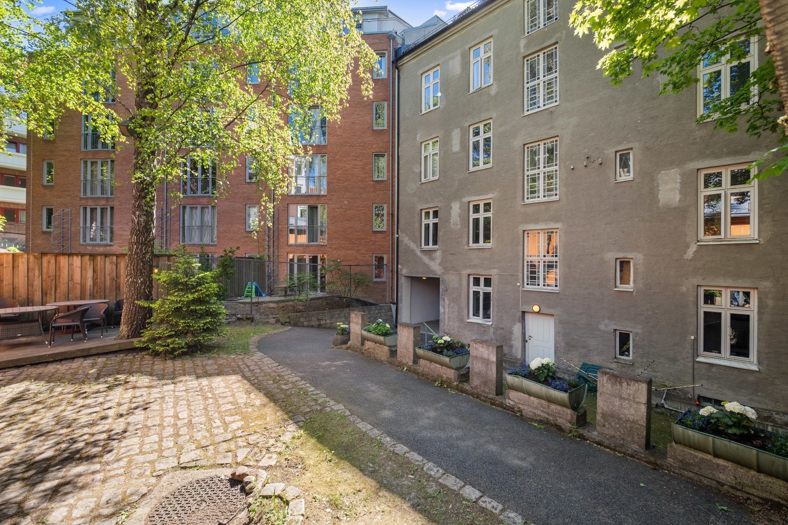 Bakgården er asfaltert og brosteinslagt, med felles terrasse, høye trær og flere blomsterkasser. Galleribilde