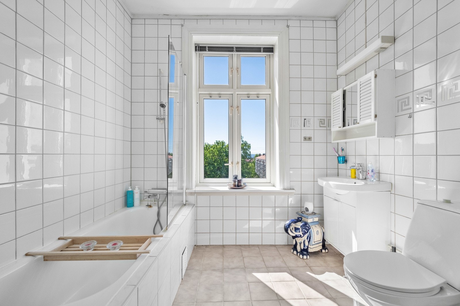 Badet er stort og lyst med krysspostvindu mot gaten, flislagte overflater og innfliset badekar med dusjvegg. Galleribilde