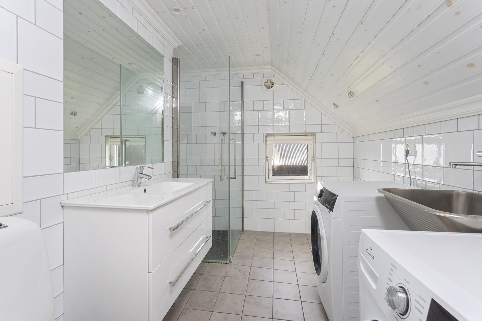 Lekkert og tidløst bad/vaskerom med flislagte overflater, hjørnedusj og opplegg for vaskemaskin - en funksjonell løsning med stilrent uttrykk. Galleribilde