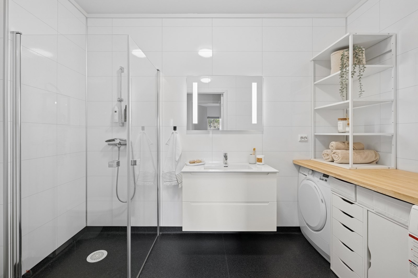 Lyst og romslig bad med opplegg for vask og tørk, servant med skap under og vegghengt speil med integrert belysning, wc og dusjhjørne. Galleribilde