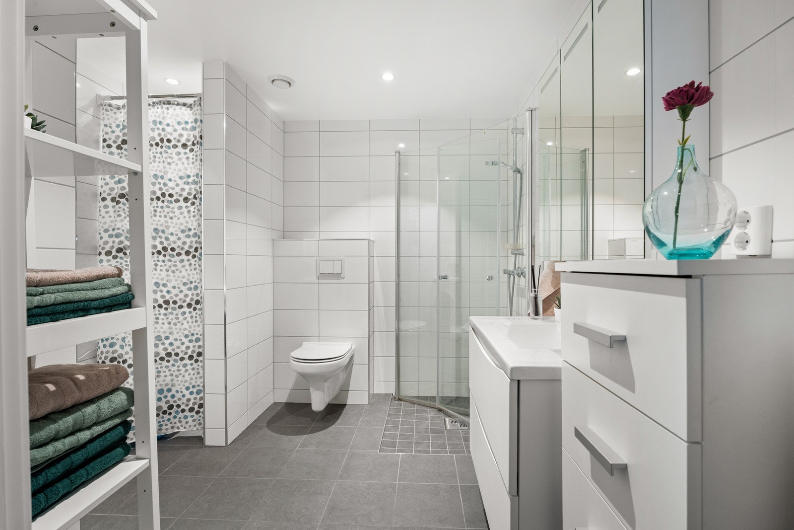 Badet fremstår som romslig, pent og velholdt, med hvite fliser på veggene, downlights i himlingen og grå fliser på gulv med gulvvarme. Galleribilde