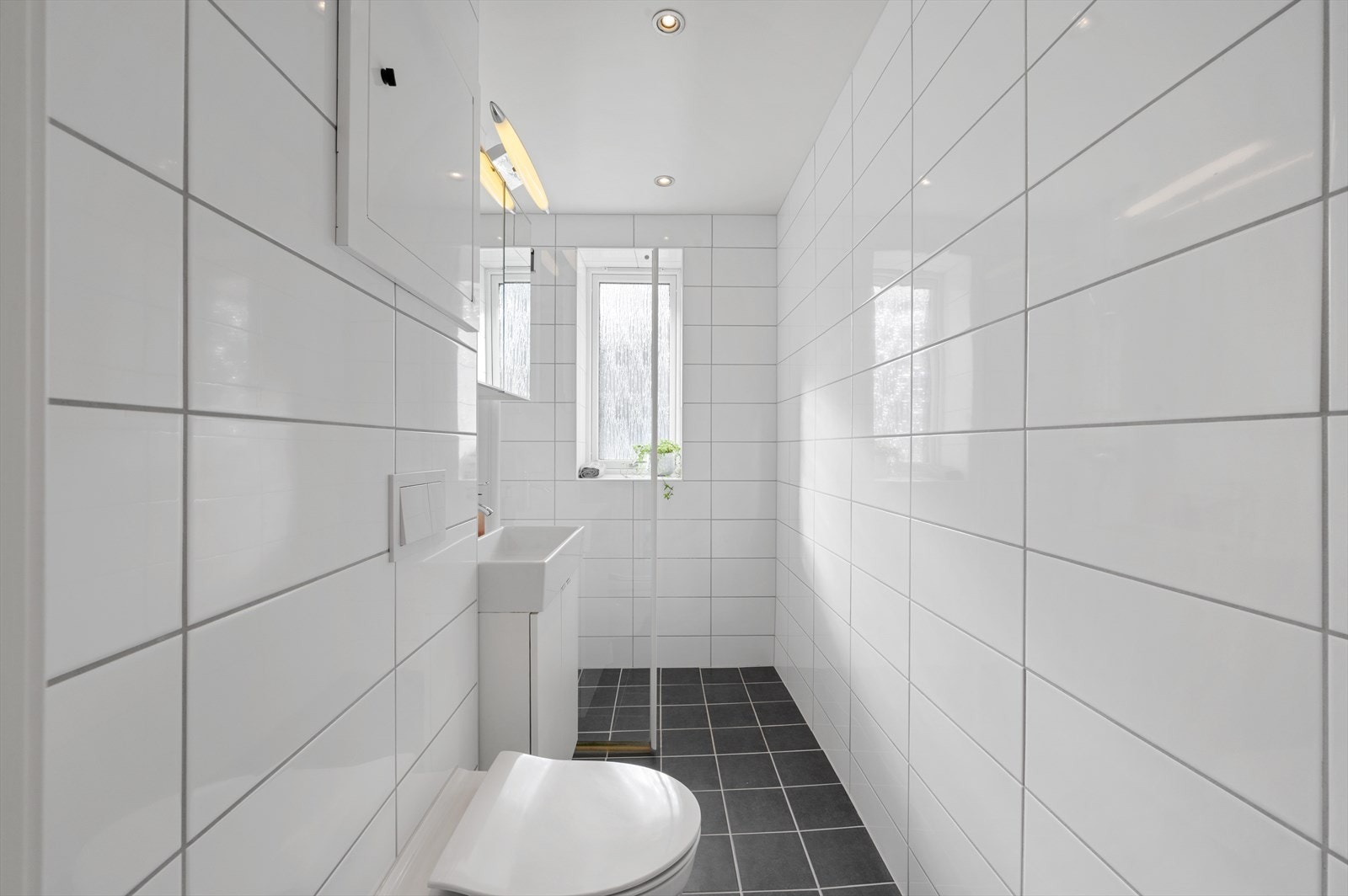 Badet er innredet med vegghengt toalett, dusjhjørne med glassdører, og servant med innredning, speilskap og belysning. Galleribilde