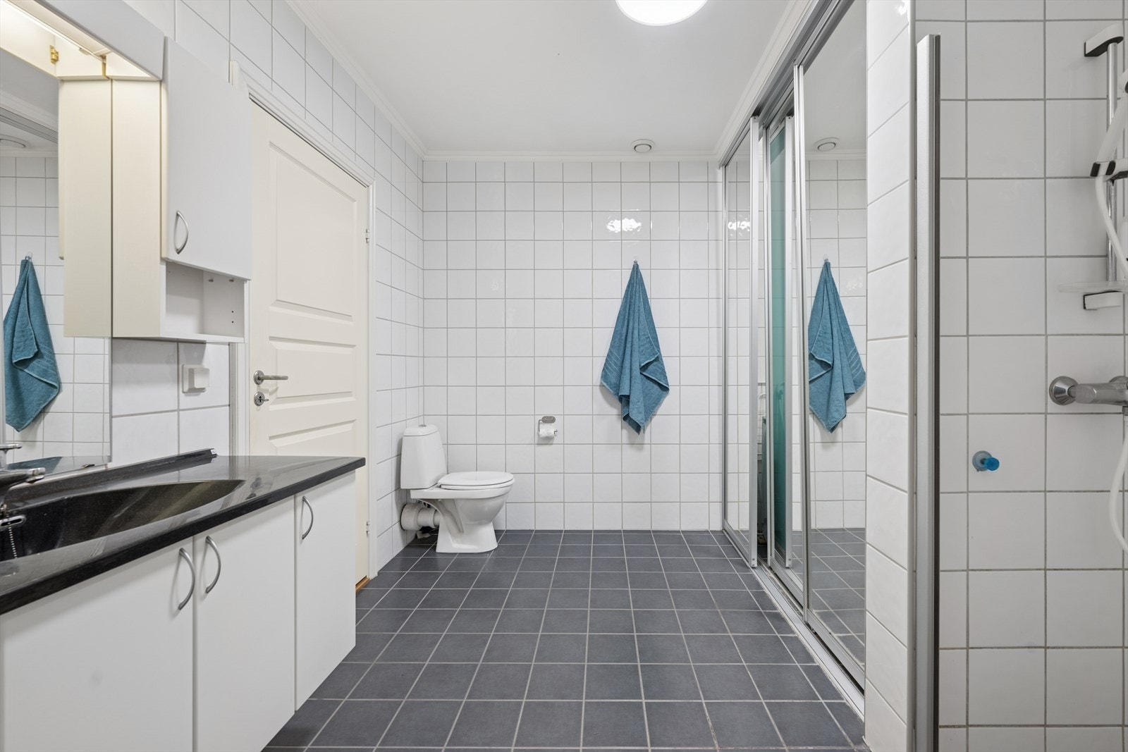 Svært romslig bad med dusjnisje, varmekabler og opplegg for vaskemaskin og tørketrommel i smart løsning bak skyvedører Galleribilde