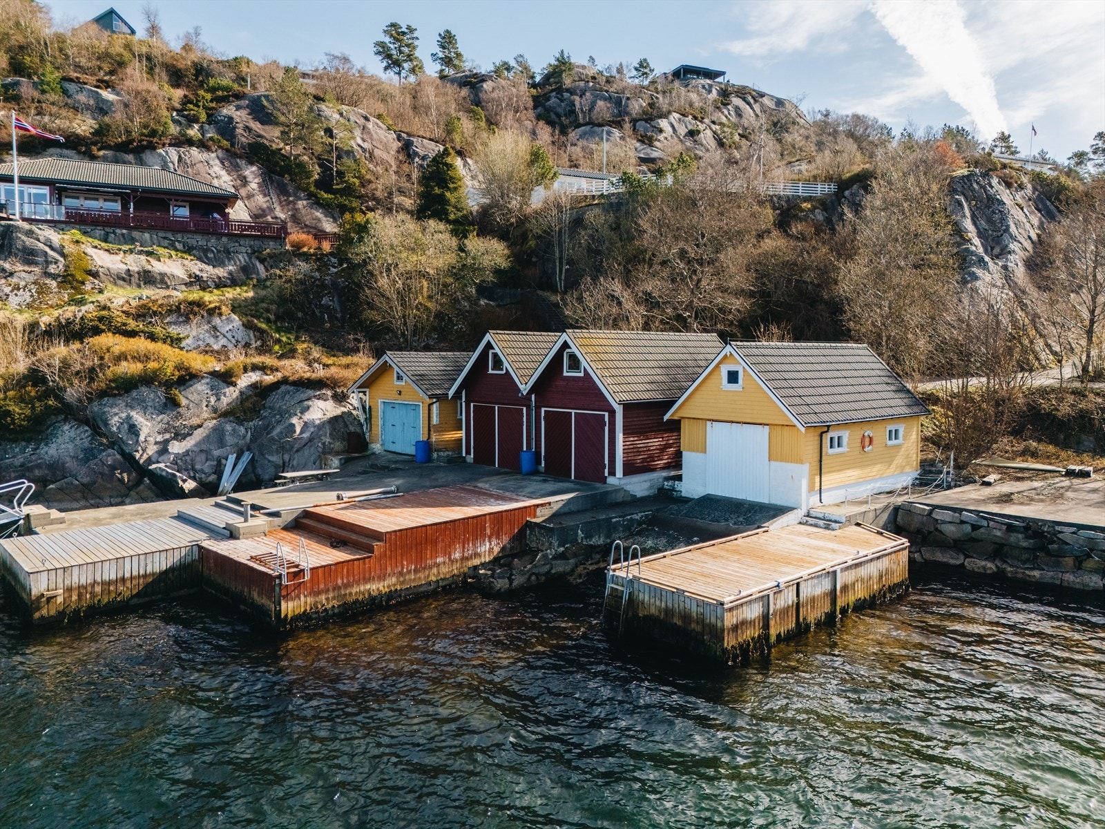 Ytterst gult naust med kai hører med. Hytten ligger pent på en hylle bak hvitt gjerde. Galleribilde
