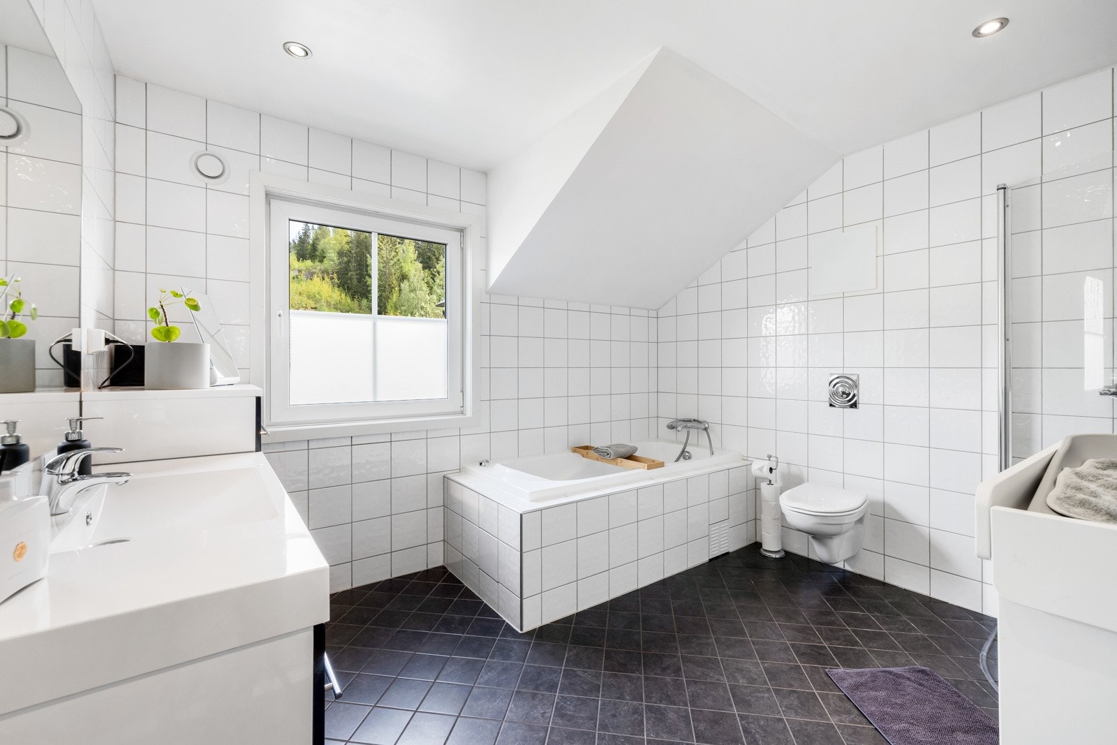 Badet har mørke gulvfliser med varmekabler, hvite veggfliser og innmurt badekar med de samme flisene. Galleribilde