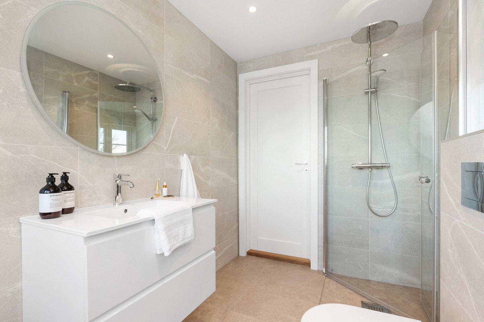 Romslig, flislagt bad som er innredet med dusj, servant i innredning med skuffer under, stort veggspeil og vegghengt toalett. Galleribilde