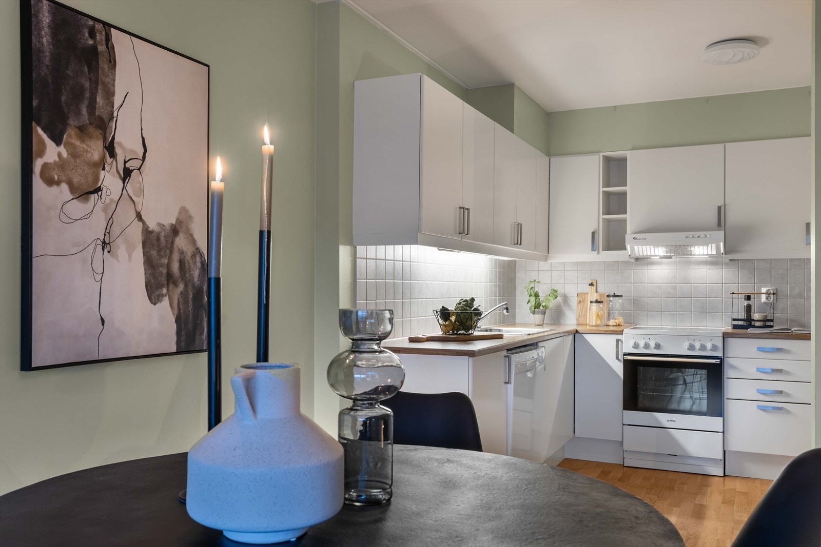 Lyst sigdal kjøkken fra byggeåret med glatte, fabrikklakkerte fronter og finert benkeplate med nedfelt kum i rustfritt stål. Fliser over benkeplaten og belysning under overskap. Galleribilde