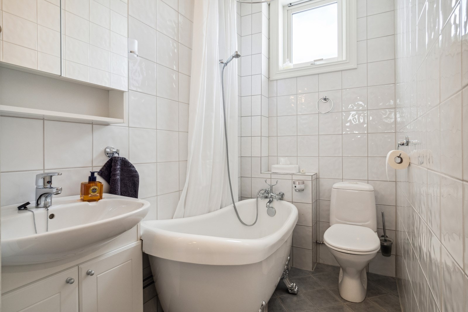 Flislagt baderom med varme i gulv, badekar, gulvstående toalett,speilskap, belysning og servant over innredning. Galleribilde