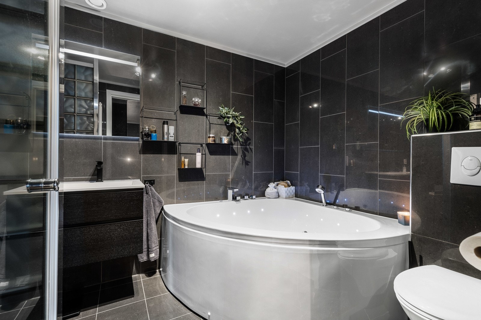 Moderne bad med mørke fliser, dusjnisje, hjørnebadekar, vegghengt toalett og servant med speil med integrert lys. Galleribilde
