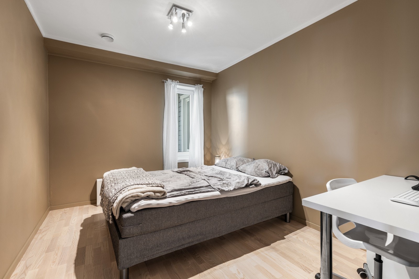 Rommet har lyse tregulv, beige vegger og et vindu med hvite gardiner som gir naturlig lysinnslipp. Galleribilde