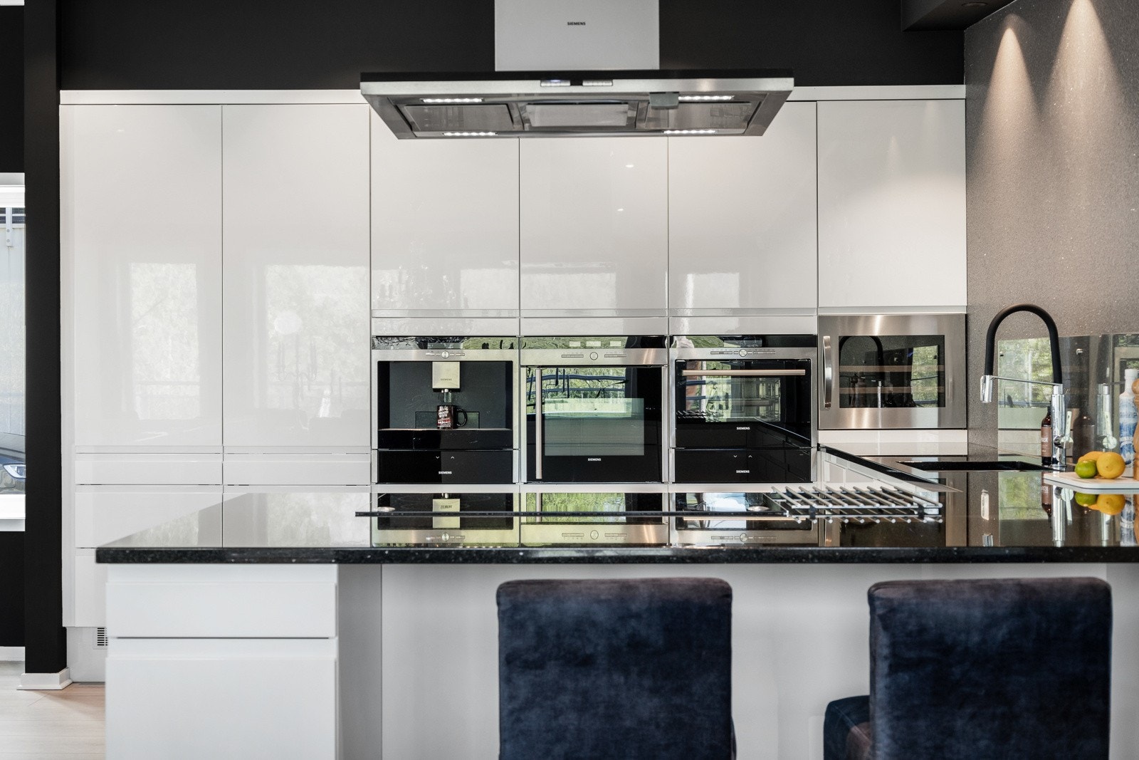 Moderne kjøkken med hvite fronter, integrerte hvitevarer og sort benkeplate. Stor kjøkkenøy med ventilator og sitteplasser. Galleribilde