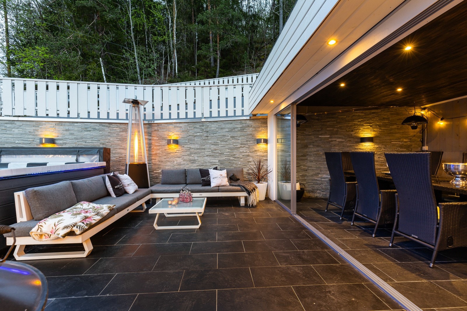 Uteområde med sittegruppe, plass til boblebad og spiseplass. Fliser på gulv, vegg med steinpanel og belysning. Skjermet av gjerde og natur. Galleribilde