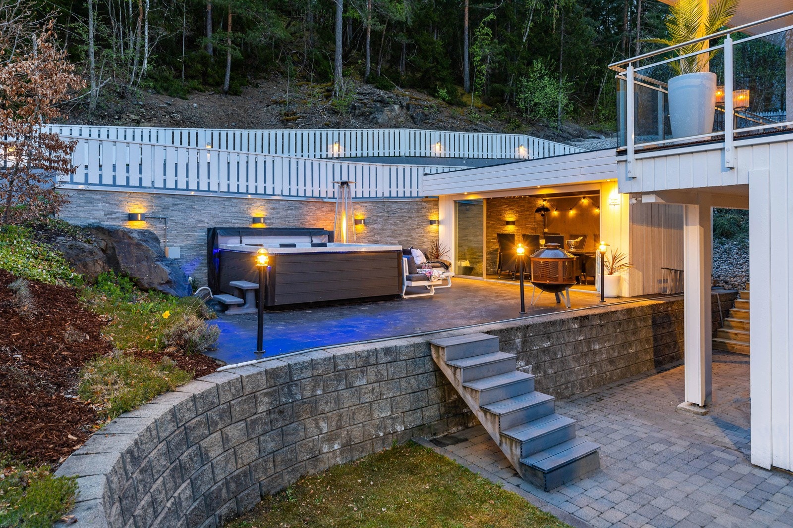 Stor, moderne uteplass med plass til boblebad, belysning og sittegruppe. Trapp til nivåer. Skjermet av skog og gjerde. Galleribilde