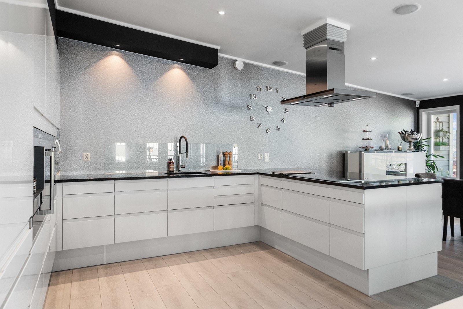 Moderne kjøkken med hvite fronter, integrerte hvitevarer og svart benkeplate. Veggklokke og lysarmaturer gir et stilrent uttrykk. Galleribilde