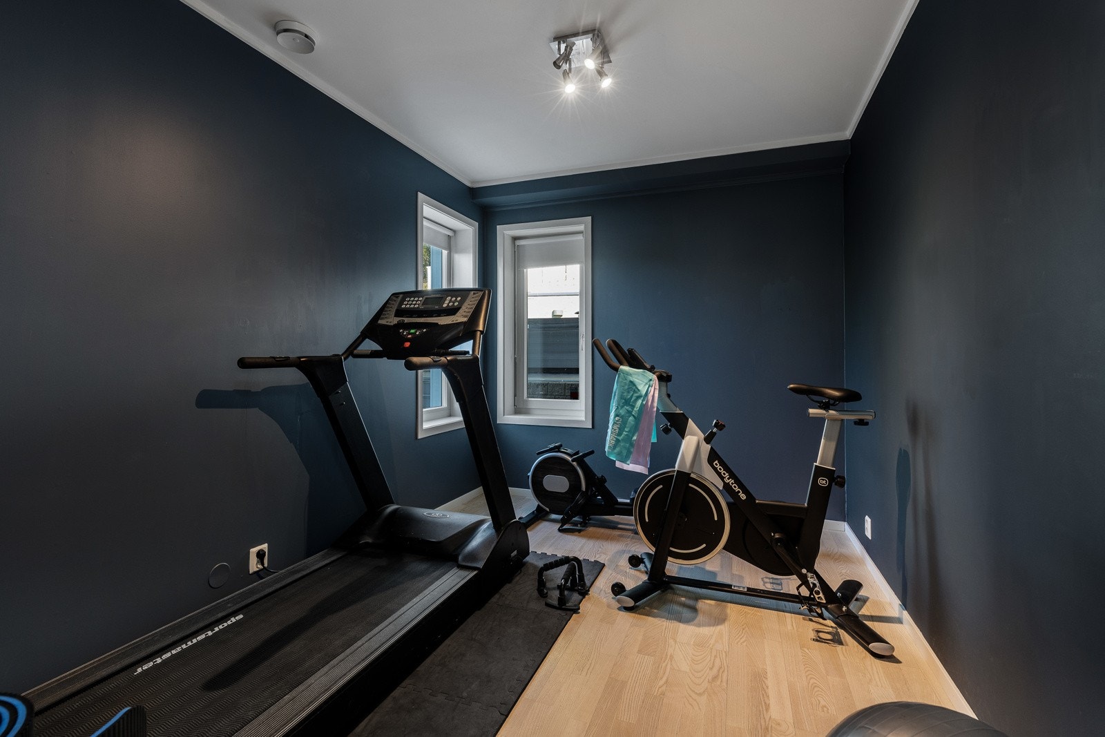 Rommet brukes som treningsrom, mørke vegger og lyst gulv. To vinduer gir naturlig lys. Galleribilde