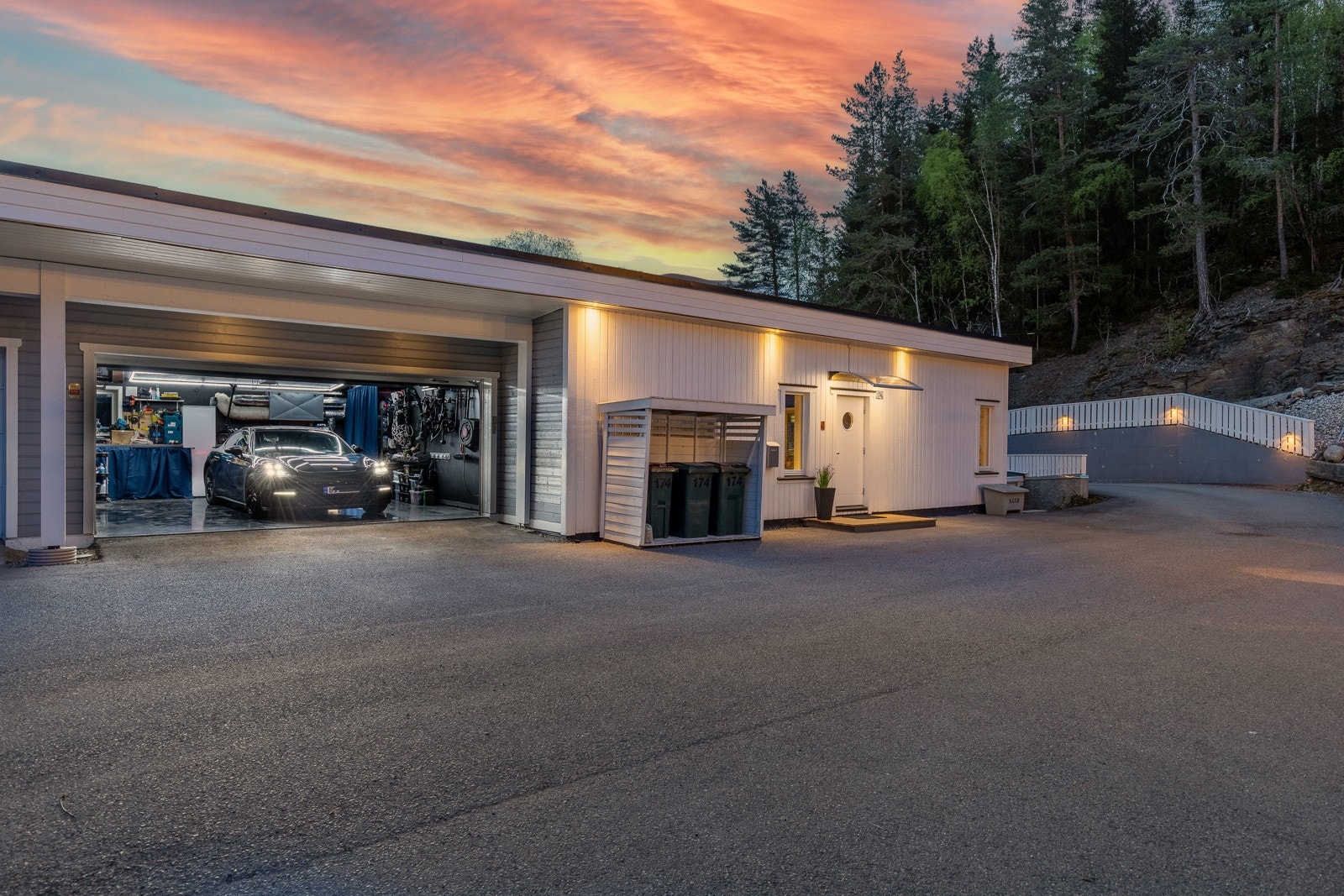 Moderne garasje med belysning, asfaltert innkjørsel og skogkledd bakgrunn. Galleribilde