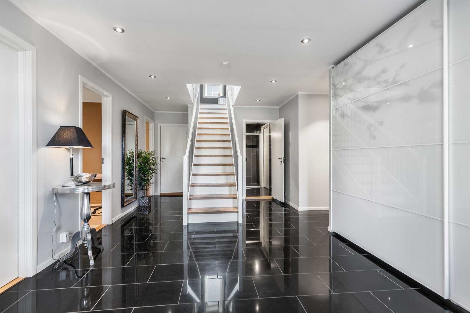 Romslig gang med svart flisgulv, hvite vegger og trapp til andre etasje. Galleribilde