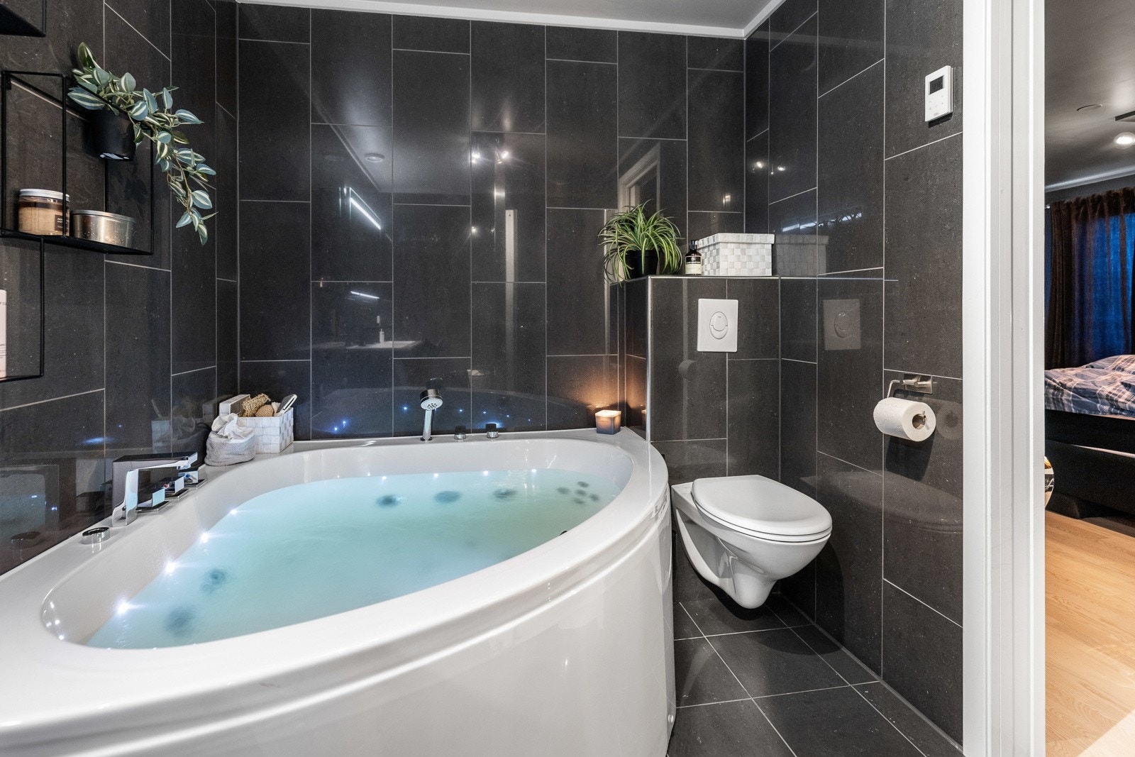 Badet har et stort hjørnebadekar og vegghengt toalett, med mørke fliser på vegger og gulv. Galleribilde