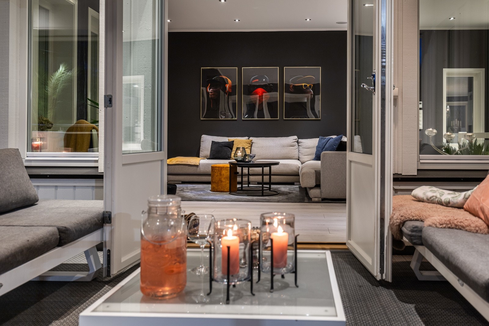 Stue med mørk vegg, lys sofa og kunst på veggen. Uteområde med sitteplasser og tente lys. Galleribilde