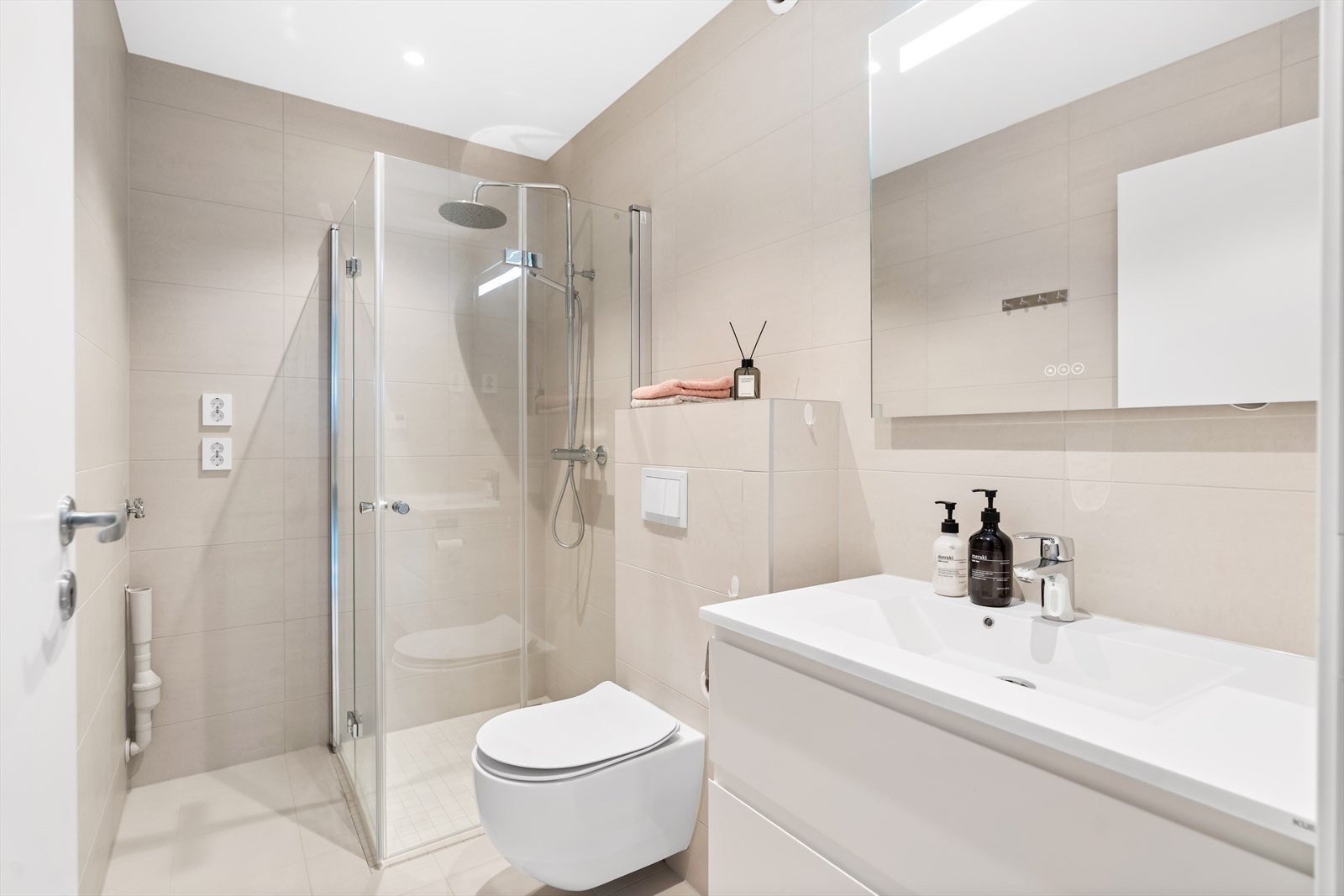 Badet i hybelen - Moderne og helfliset bad med vegghengt toalett, dusjhjørne med glassdører, servant med skuffer og opplegg for vaskemaskin. Galleribilde