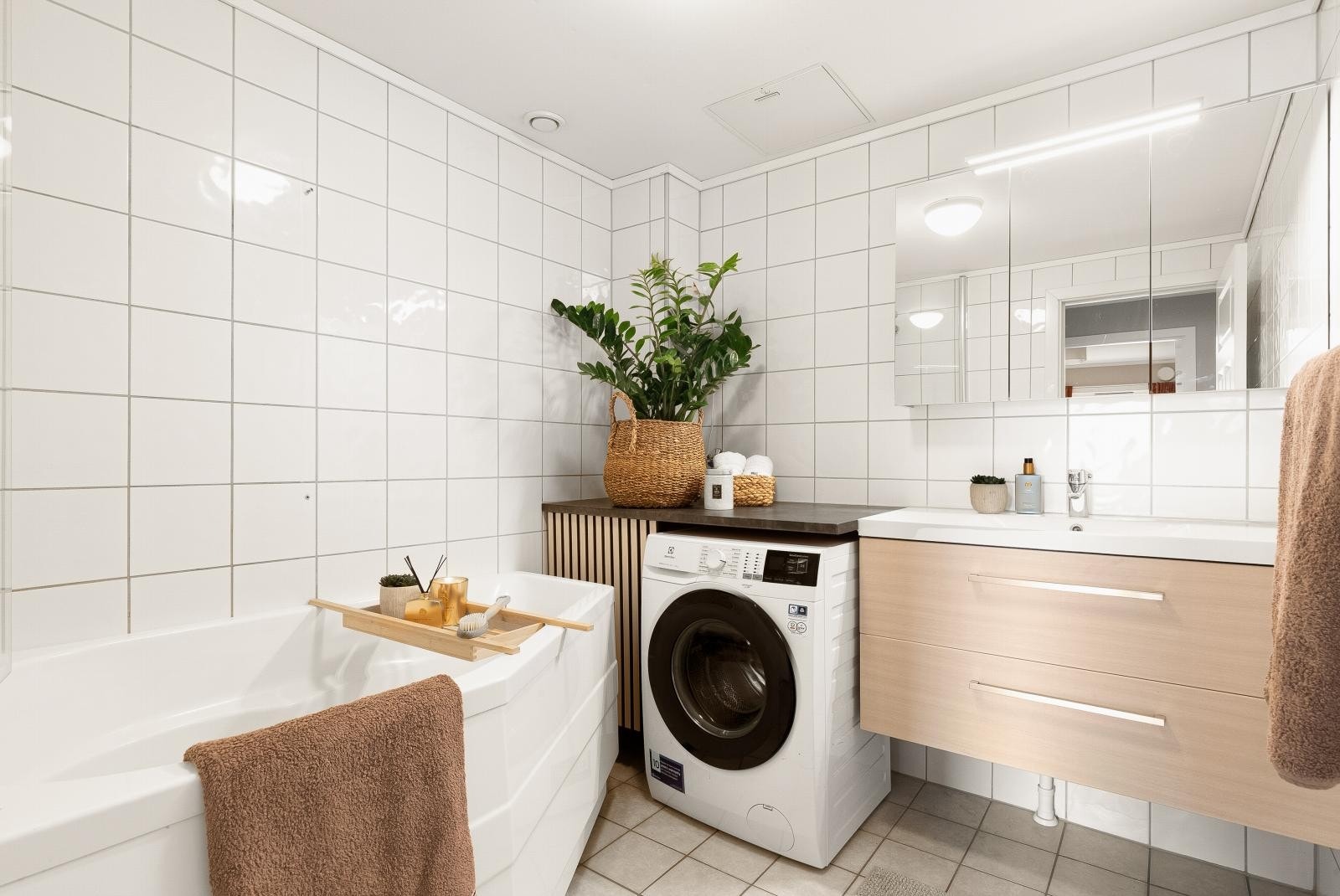 Lyst og pent bad med badekar, baderomsinnredning med glatte fronter og opplegg for vaskemaskin. Rommet har lyse fliser og gulvvarme. Galleribilde