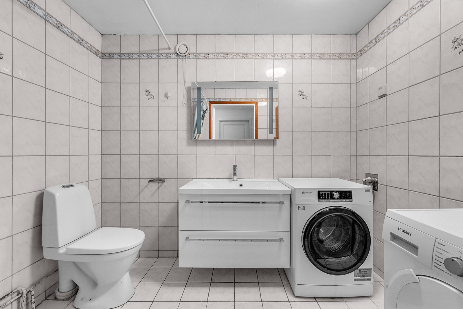 Praktisk bad/vaskerom med flislagte gulv og vegger samt malt himling. Rommet har klosett, heldekkende servant med underskap(oppgradert i 2022), speil med belysning og dusjhjørne med dusjforheng. Det er opplegg og plass for både vaskemaskin og tørketrommel Galleribilde