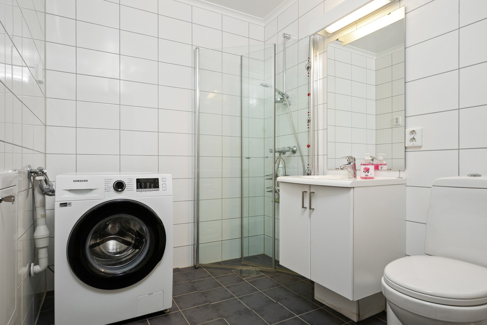 Badet har mørke fliser på gulvet, lyse fliser på veggene, gulvvarme og opplegg for vaskemaskin. Galleribilde